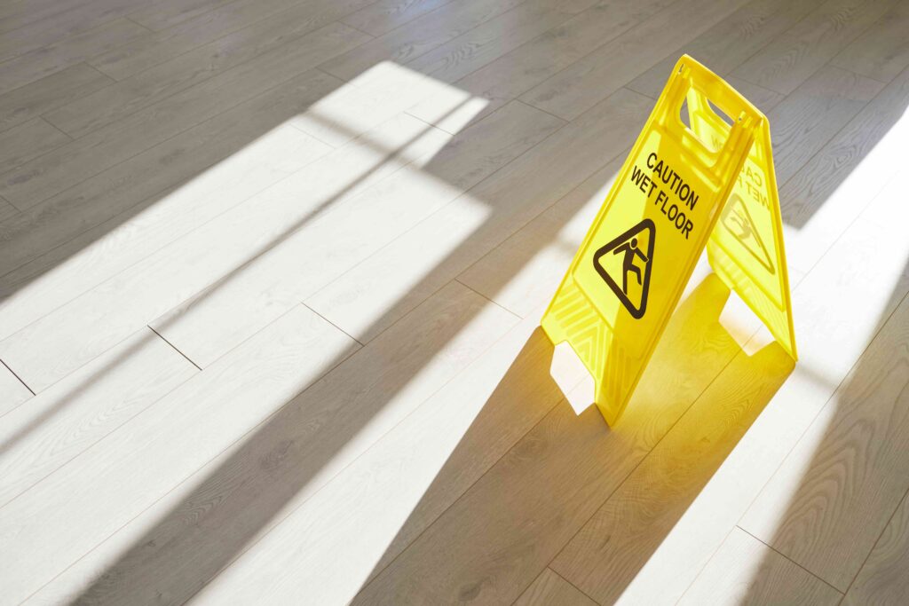 Yellow caution wet floor sign on hardwood floor with sunlight and shadows, warning of slip and fall hazard.