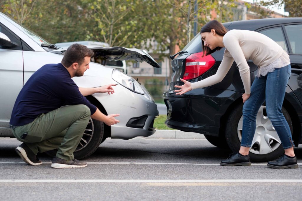 Man and woman inspecting damage after a rear-end car accident, showing dented bumper and open hood in parking lot.
